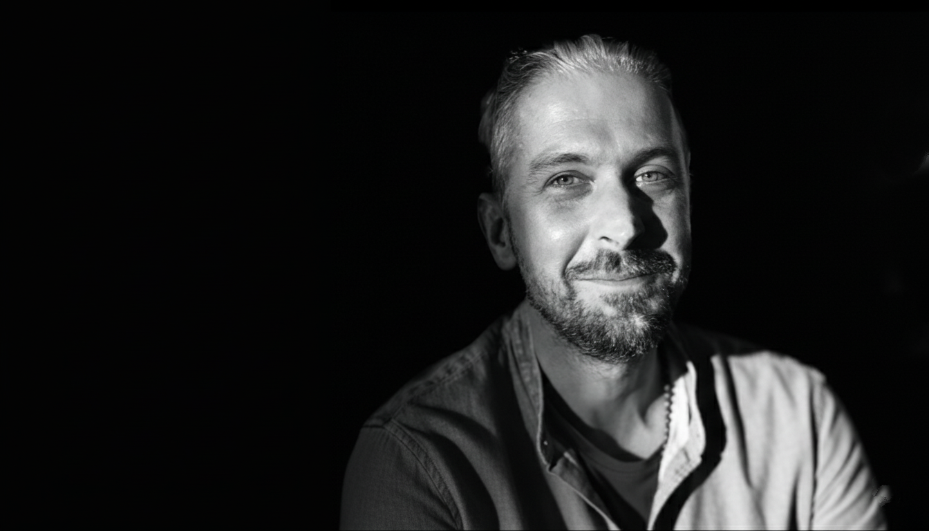 Black and white image of Alex Crockett smiling with a dark background and light illuminating one side of him.